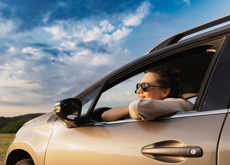 A smiling driver with sunglasses in a parked car smiling while leaning out the window in a open landscape.