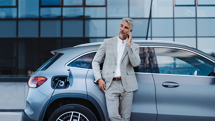 A man in a suit is leaning against a car while talking on a cell phone