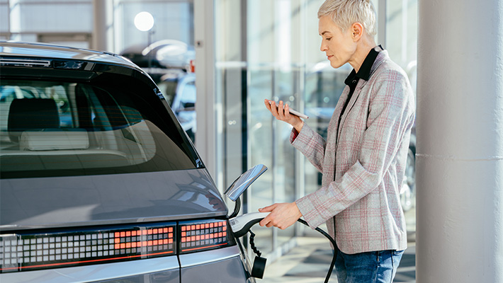 A woman is charging her electric car at a charging station while looking at her phone