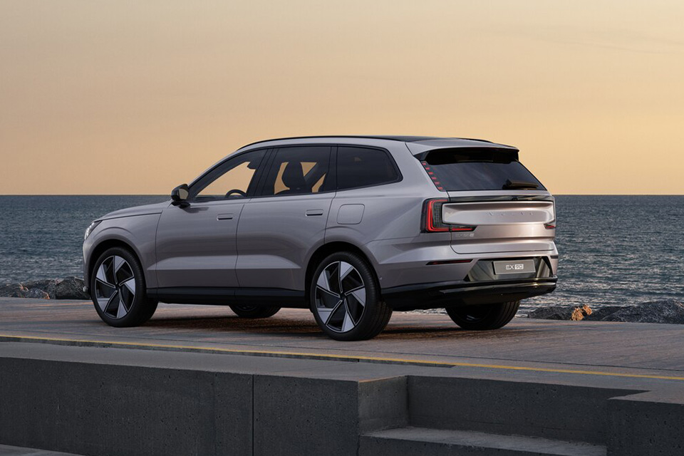 A sleek, silver Volvo EX90 is parked by the water at dusk