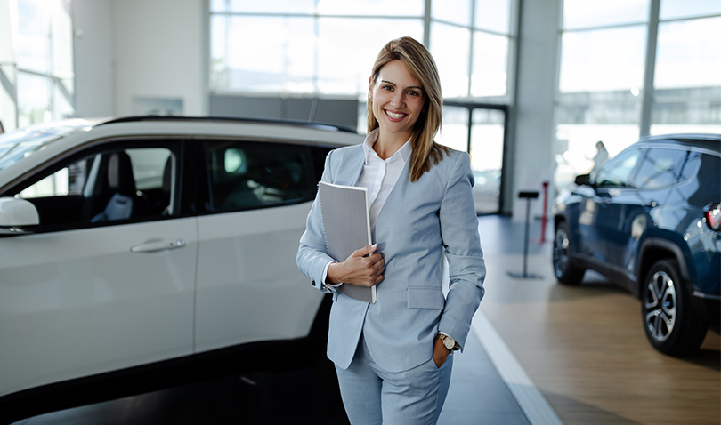 A woman in a suit is standing in front of a car in a showroom holding a tablet